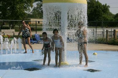 Splash pad / waterpark photo with kids under a mushroom fountain — shows the campground’s pool/water play facility mentioned on the site.
