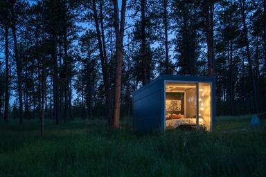 Twilight/night shot of a small modern cabin with interior lights visible through large windows set in the woods — offers an atmospheric view of the property’s lodging experience.