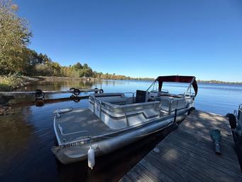 Pontoon boat docked on a wooden pier next to a lake under a clear, blue sky.