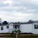 Wide image of a mobile home with landscaped foreground and a lake/trees in the background — good for showing rental unit appearance and grounds (source: Rentals wrapper, rentimages/10.jpg).