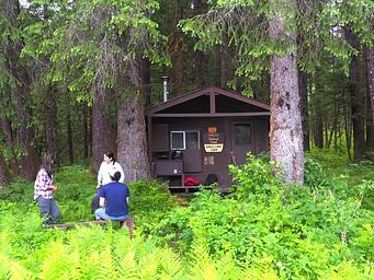 Three people outside brown cabin with trees and ferns
