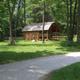 Wooded campsite with a small log cabin/shelter, picnic tables and gravel access road — matches hero/background images on the campground page showing on-site cabins and campsites.