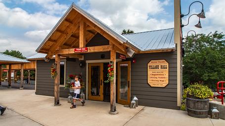 Exterior of a timber-framed building labeled 'Village Hall' with entrance plaza — shows on-site facilities and visitor amenities.