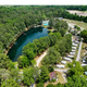 Clear aerial overview showing the pond, surrounding tree line, pool area, RV rows and roads — good overall site layout shot for the property gallery (matches homepage drone images).