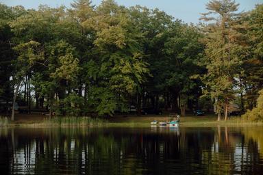Wide lakeshore view across the water showing treeline, shoreline with small dock and camping area — good overall property overview. Sourced from the Our Campground page (EPLC-23) representing the campground’s lake frontage.