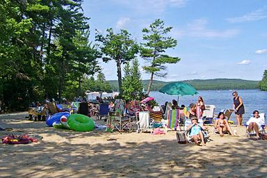 Beach scene with campers, chairs and lake in background — illustrates the campground's sandy swimming area and family recreation on Long Lake.