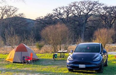 Tent campsite with tent, picnic table and vehicle visible on grassy pad near trees and riverbank — represents tent camping sites and setting on the property.
