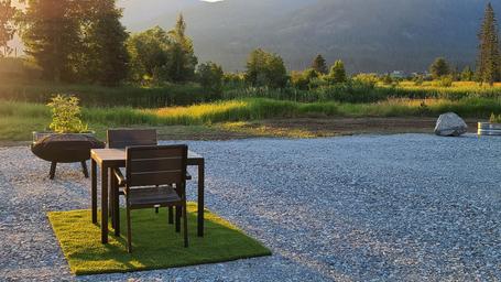 Sunlit campsite vignette with table, chairs and a fire ring on gravel pad looking toward the lake/landscape — shows guest seating/amenity detail used in the property's listing.