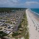 Aerial view of a beach with umbrellas and people on the sand, beside an oceanfront campground filled with cars and RVs, with a forest in the background and the ocean on the right.