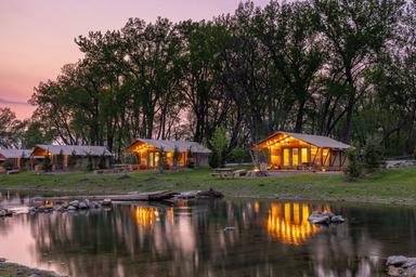 Twilight lakeside shot of illuminated glamping cabin/tent reflecting in the pond — strong scenic image showcasing waterfront accommodations and ambiance (used on homepage).