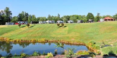 Panoramic hillside shot showing a line of RVs parked above a pond with landscaping and a covered picnic shelter — demonstrates campsite layout and grounds (from Amenities page).