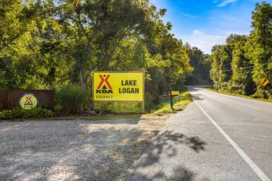 Entrance roadway with prominent 'Lake Logan KOA' sign and surrounding trees — establishes property identity and shows the campground entrance (used as a background/intro image for the campground page).