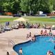 Clear daytime photo of the on-site swimming pool with lounge chairs, people using the pool and campground structures (kayak/rv area) visible in background — useful to show the facility and pool area.