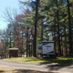 Clear on-site photo showing an RV parked among tall pine trees with campsite infrastructure (road, small shed/sign). Supports depiction of RV pitches and wooded campground setting on the property's homepage.