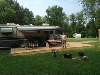Large RV parked at a long pull-through site with a wooden deck, chairs and fire ring — illustrates the campground’s RV/Park Model accommodations and site setup shown in the ‘ways to stay’ photos.