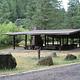 Large picnic shelter with stone fireplace and chimney and boulders lining a gravel road in front of fir forest and shrubs.