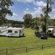 View of RV sites on mowed grass with rigs and vehicles and trees in the background — demonstrates RV camping layout and site spacing.