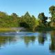 Clear wide photo of the property pond with fountain and tree-lined shore — used as the site’s main header/slider image (page background/slider), showing on-site scenery and water feature.