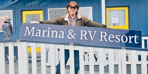 Man in cowboy hat holding a Marina &amp; RV Resort sign outside a blue building.