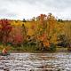 A view from the river facing the land. A canoe with two paddlers paddle towards a forest with vibrant red and yellow leaves on the canopies of the trees.