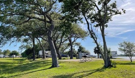 A grassy, treeâshaded area in an RV park with several RVs parked on paved pads in the background. A picnic table and grill sit near the water, and the sky is clear with scattered clouds.