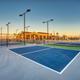 Empty outdoor pickleball courts with blue and green surfaces, surrounded by fencing and tall lights, near a modern building and a playground under a clear sky at sunset.