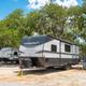 RV site with travel trailer parked on a defined pad among trees; shows typical RV pitch surface, hookups and surrounding landscape.
