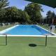 Rectangular outdoor swimming pool with clear water, surrounded by green deck, tables, chairs, and trees in the background under a sunny sky.
