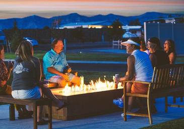 Evening photo of guests around a built-in firepit with RVs and mountains in background — shows communal amenity and evening atmosphere on the resort grounds (used in amenities section).
