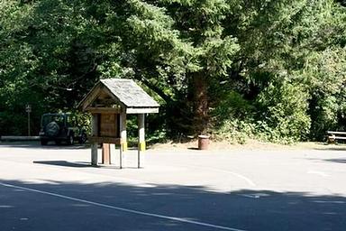 Paved entry with kiosk and fee tube in front of parking area and large evergreen shrub tree.