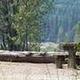 Picnic table in a flat gravel area with bushes and log parking barrier, river valley and conifer covered hills in background.