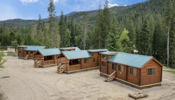 A row of cozy wooden cabins with green metal roofs sits in a forest clearing, mountains and tall trees in the background, under a blue sky.