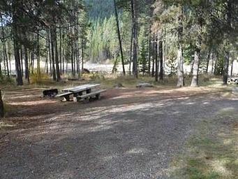 Flat gravel parking area next to picnic table and fire ring in pine forest with lake in the background.