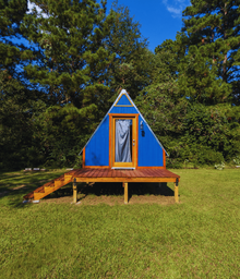Exterior of a small blue A‑frame cabin on a grassy site with trees behind — clear image of one of the on‑site lodging units featured on the property pages.