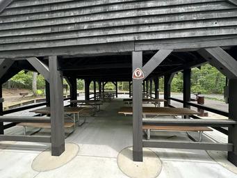 Eight picnic tables sit on a concrete pad under a wooden open sided picnic shelter