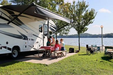 People relaxing beside a white RV under an awning by a lakeside campsite