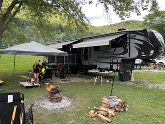 Close-up campsite scene with a large fifth-wheel RV, awning, fire ring and grassy pitch area — useful for showing typical RV site setup and amenities on the campground grounds.