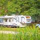 Clear daytime photo of an RV site with a trailer, parking, and natural screening by trees with a pond/reeds in foreground — shows typical RV pitch on the property (image appears on the campground’s Arrowhead RV Resort listing page).