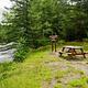 A gravel and grassy campsite to the right of a river. The campsite has a wooden picnic table, a brown wooden sign, and a trail that leads into the woods with tall dense green trees.