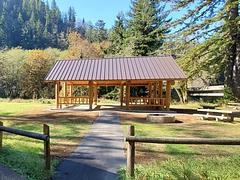 Large flat field.  Tents and picnic tables in front of a road and large conifer treein the back, fire ring and picnic table behind single pole fence in foreground.