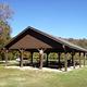Front view of the covered pavilion with picnic tables surrounded by a grassy field. Green trees beginning to change colors are visible in the bakckground.