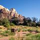View along the walk-in path to a campsite in Zion Canyon, featuring a picnic table, fire ring, and food storage locker among low vegetation, with rugged sheer sandstone cliffs rising dramatically in the background.
