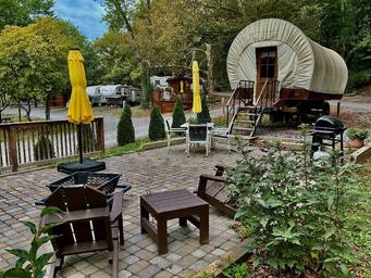 Covered-wagon style lodging and a furnished patio with chairs and umbrellas — shows a unique on-site lodging option and landscaped guest area from the campground’s imagery.