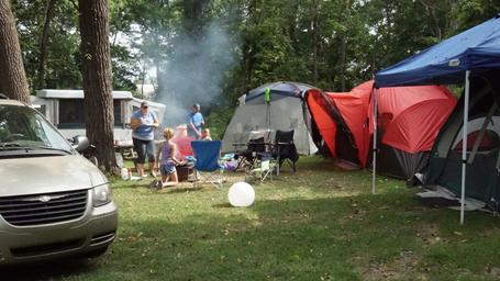 Active tent campsite scene with tents, chairs and a vehicle in a wooded site — useful depiction of tent camping options; originates from the site's campsites imagery.