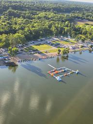 Aerial view of Lazy L Lake Campground showing RV sites, the sandy swimming beach lined with kayaks and floats, and the inflatable aqua park on the 75-acre lake