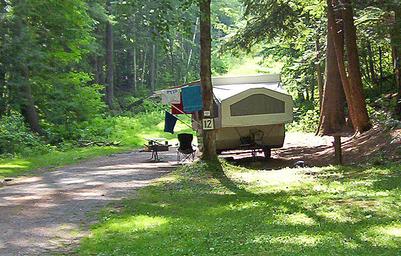Campsite image with pop-up camper, site marker (#12), picnic table and access road — illustrates site amenities and grassy/wooded terrain.