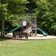 A small playground nestled in the trees and surrounded by manicured grass.