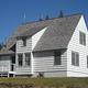 White house with dormer roof on a grassy hill with conifer trees.