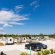 A sunny RV park with several parked trailers and trucks on gravel lots, surrounded by trees and under a partly cloudy blue sky.
