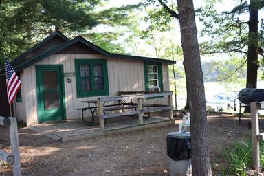 Small cabin with porch, picnic table and lake glimpses beyond — another on-site cabin view that illustrates campsite layout and nearby shoreline (from the resort site).